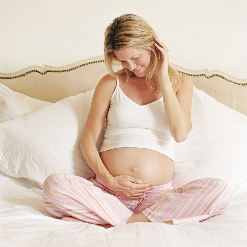 Smiling pregnant woman touches her belly as she sits on a bed looking comfortable